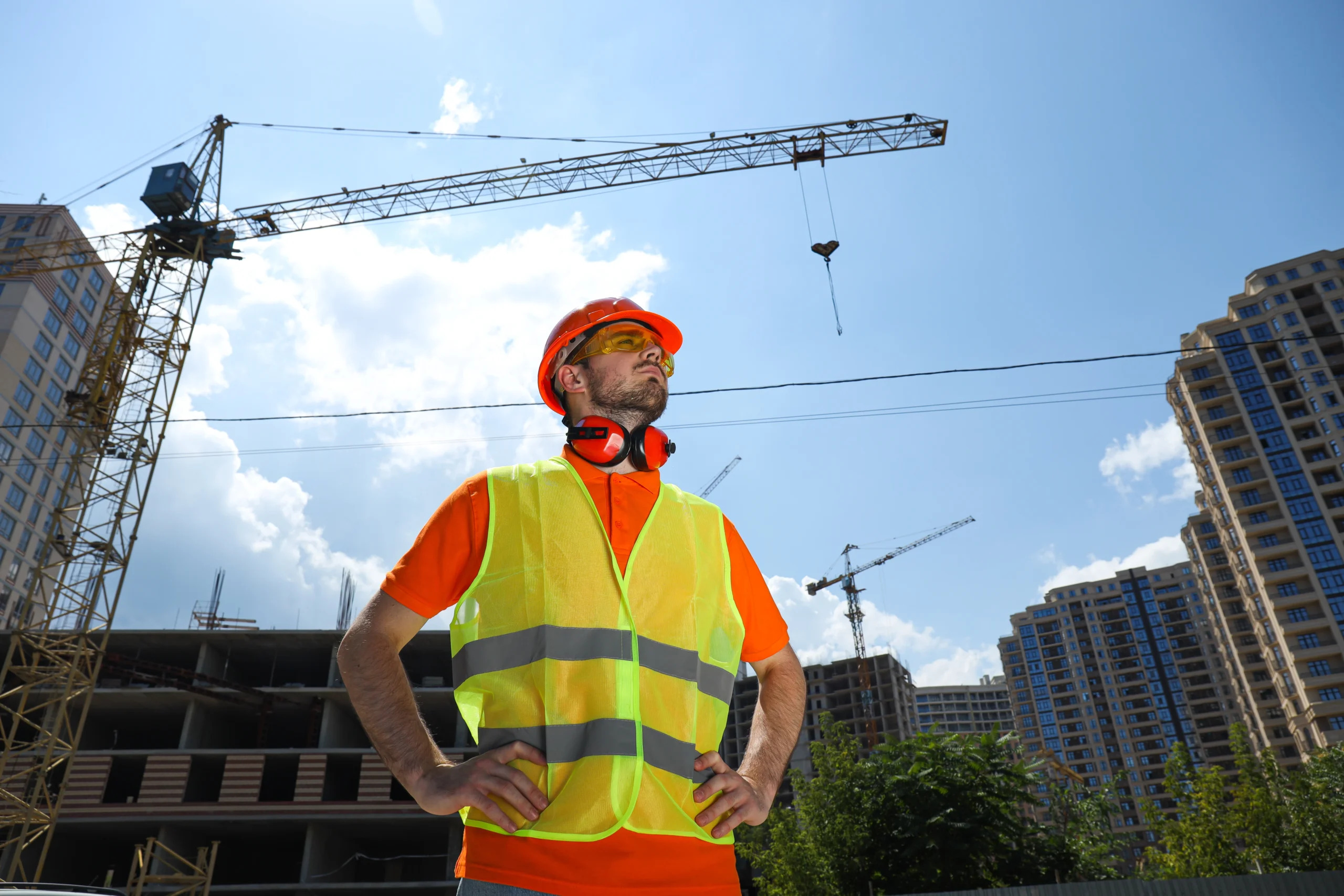 Construction worker wearing a hard hat and high-visibility vest at a building site with tower cranes.